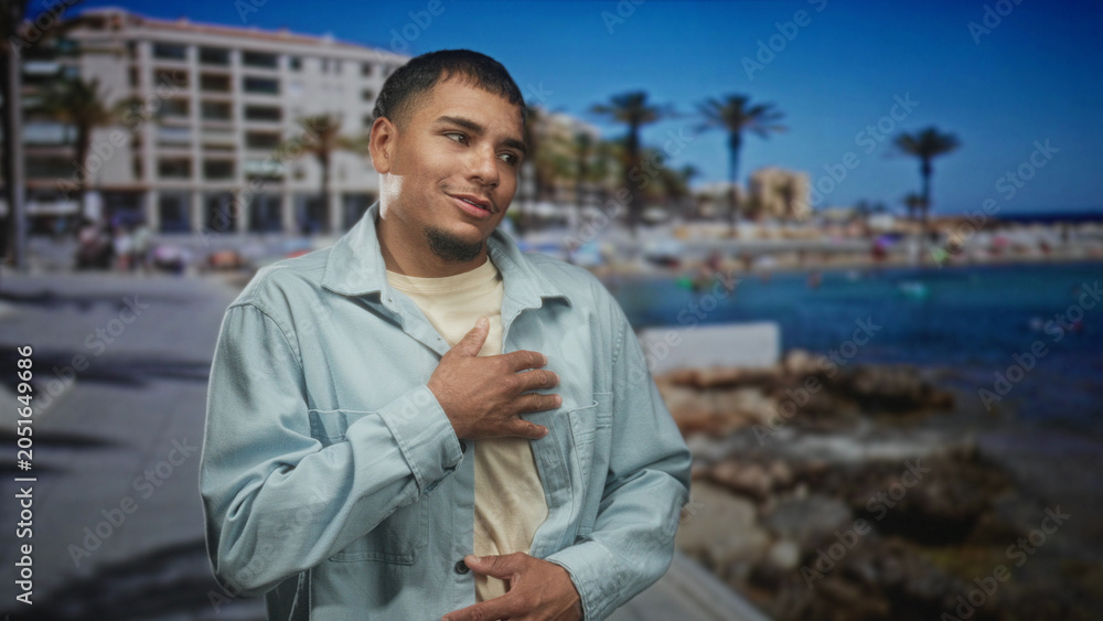 Fototapeta premium Man with hand on chest in a light jacket by palm trees and buildings on a street, smiling and leaning forward; sincere gratitude.