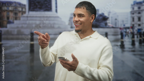 Φωτογραφία Man tapping smartphone with both hands and briefly pointing finger while standing on a wet street near a monument; curiosity