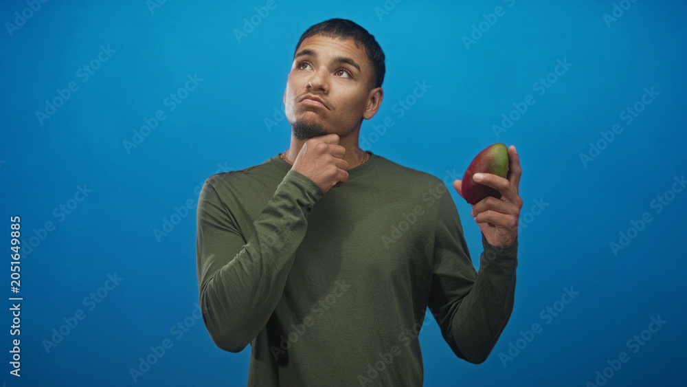 Fototapeta premium Man holding mango with hand on chin in studio, inspecting the fruit and weighing ripeness and flavor options; thoughtful choice.