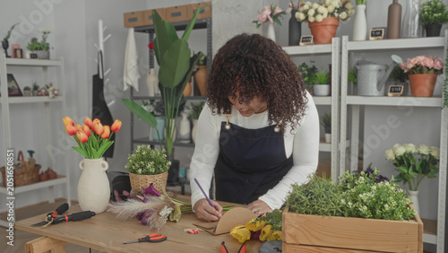 Fotografie Woman florist writes a label on kraft tag while leaning over tulips, scissors and potted greens in studio; creative serenity