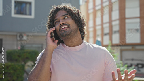 Fotografie Man holding smartphone to ear, smiling and gesturing on a sunny street in front of buildings; friendly conversation cheerful