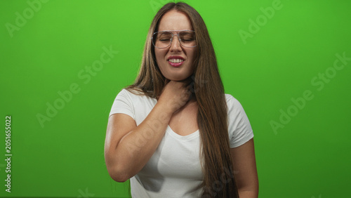Photos Young caucasian woman clutching her neck and grimacing in a green screen studio with glasses and white tee; throat pain discomfort