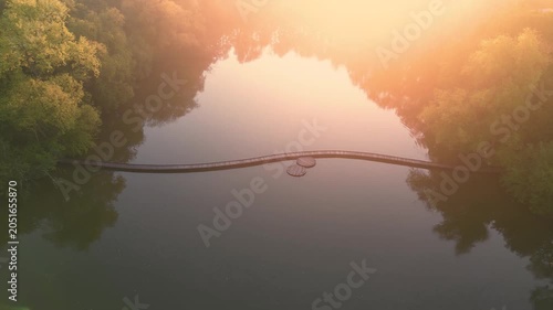 Sunset casts light on a lake with a wooden path. Trees surround the water. The wooden path leads across the lake, inviting exploration during dusk hour. Aerial drone footage