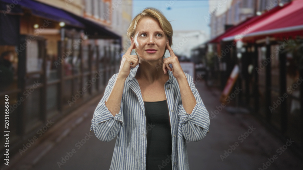 Fototapeta premium Woman in a striped shirt gesturing with open hands toward camera on a bustling street with sidewalk cafes, awnings and outdoor tables; thinking.