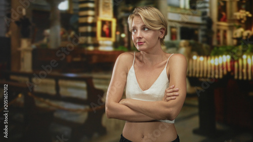 Bild auf Leinwand Caucasian woman with arms crossed revealing navel and wearing a white camisole inside a church interior with lit candles, wooden pews and paintings; contemplation thoughtful