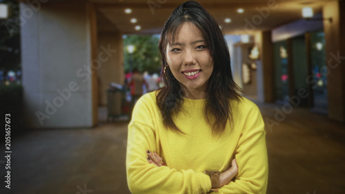 Tableau sur toile Young woman in yellow sweater with arms crossed and earrings smiling at camera in building entrance; confidence warmth