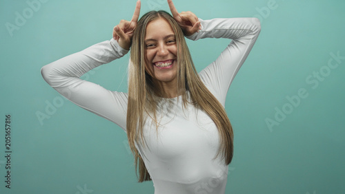 Foto Young woman smiling and making horns gesture with fingers to head in studio wearing white long sleeve top against teal backdrop; playful mischief