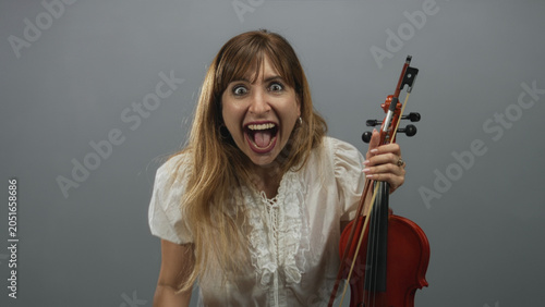 Woman holding violin and bow with open mouth shouting pose in gray studio wearing white blouse; raw excitement.