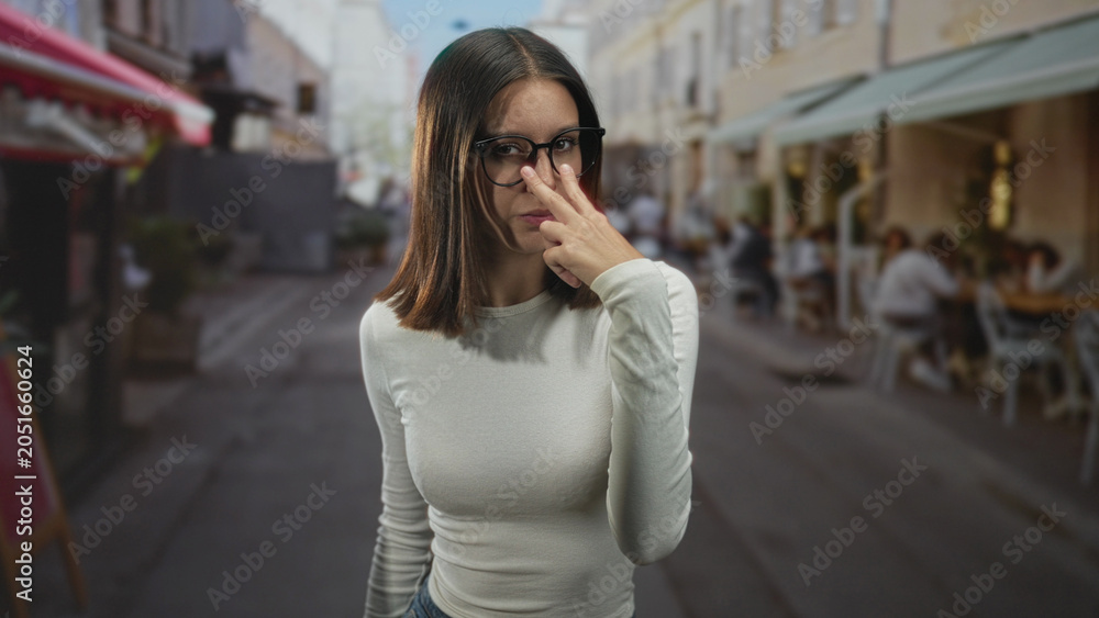 Fototapeta premium Woman wearing glasses points finger toward camera while standing near outdoor cafe tables on a street; confidence challenge.