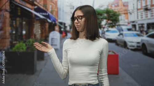 Tableau sur toile Woman pointing index finger near face in front of a storefront on a busy street, wearing glasses and a white long sleeve shirt with a dismissive expression; annoyance dismissal