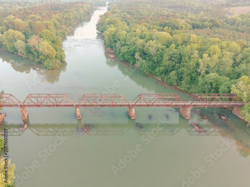 Rusty bridge spans calm, green river. Lush trees line both riverbanks. Reflections ripple gently below. Industrial decay meets natural beauty. Ideal for travel, nature, or nostalgia themes