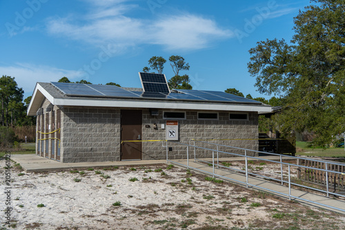 Solar panels cover the roof of a stone building. Wooden door stands under a small overhang. Yellow tape marks the building's perimeter. Trees rise behind, under a blue sky