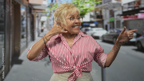 Fotografie Senior woman making phone gesture with hand and pointing finger in a lively urban street scene; playful greeting joy