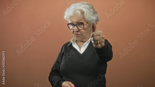 Tableau sur toile Elderly woman with white hair and colorful eyeglasses extends thumb down gesture toward camera in studio; disapproval judgment