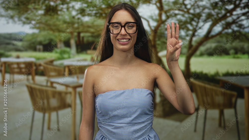 Naklejka premium Woman wearing glasses and strapless dress raises hand showing three fingers at a restaurant terrace building; cheerful greeting.