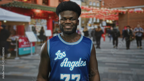 Foto Man smiling in blue los angeles jersey on urban street with market stalls and lanterns visible; casual joy