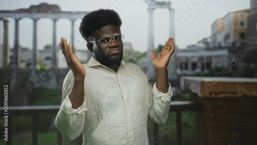 Obraz na płótnie Man with hands raised palms up shrugging at roman building among ancient ruins, wearing glasses and linen shirt; confusion uncertainty