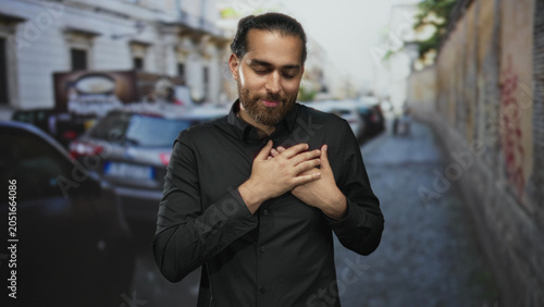 Fotografie Man with hands over chest on a city street wearing black shirt, eyes closed in a thankful gesture; gratitude reflection