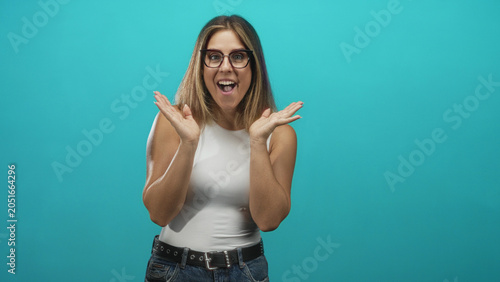 Fotografie Woman with raised hands and open mouth showing surprise in studio with turquoise backdrop, white sleeveless top and jeans; celebration excitement