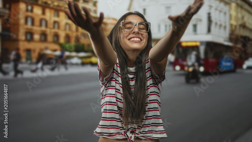 Fotografie Woman with glasses and striped shirt beckons with hands on a busy city street beside a red bus and classical building; playful