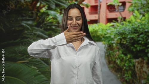 Canvas Print Woman in white shirt with hand over mouth and raised palm near face on street beside a building and lush tropical plants; shy smile introspection