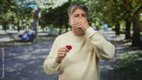 Foto Man holding small red heart, hand pressed to mouth and looking distressed on a tree lined street in a park; heartache concern