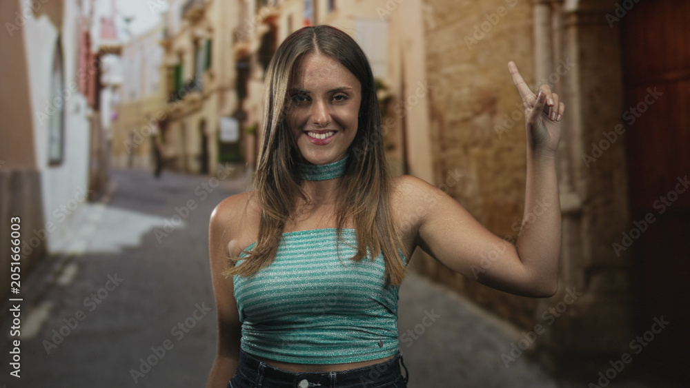 Naklejka premium Woman raises bare arm and points finger upward on a narrow street wearing turquoise crop top and choker; effortless confidence.