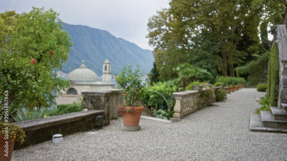 Obraz premium Blurred terrace scene with potted plants, gravel path and distant domed building in soft defocus, garden; background backdrop copyspace calm.