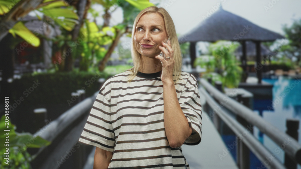 Fototapeta premium Woman holding smartphone to ear on resort pool walkway near gazebo building during phone call; quiet contemplation.