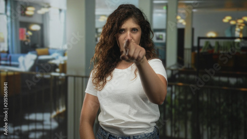 Bilde på lerret Woman points finger at camera in hotel lobby with stern gaze and extended arm, white t shirt visible; accusation