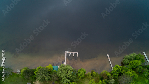 Foto A direct overhead drone photograph looking down at a large E-shaped wooden boat dock and smaller piers along a clear sandy shoreline, transitioning into deep dark water under a moody spring sky