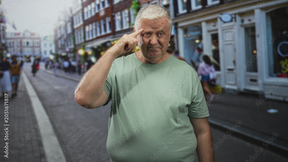 Fototapeta premium Man points finger to temple on a busy street near storefronts and sidewalk, wearing a green tshirt and standing center frame; thoughtful reflection.