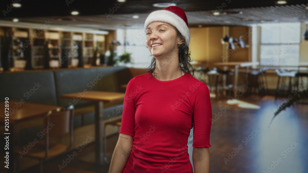 Fototapeta premium Woman wearing santa hat smiling and looking up inside a building cafe with wooden tables and chairs; cheerful holiday.