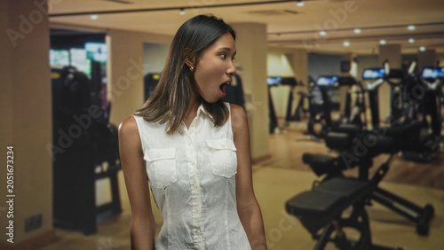 Photos Thai woman yawning with open mouth beside gym bench, head turned to the side and arms relaxed; fatigue rest