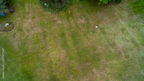 A high-angle top-down drone photograph showing a single person standing in a massive, expansive green and brown grassy lawn field with trees and a small garden feature visible along the margins.