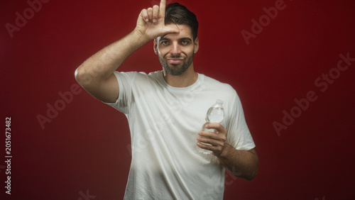 Bild auf Leinwand Hispanic man with beard in white t shirt holding a water bottle and making an l on his forehead while alternating pointing gestures in studio; playful tease