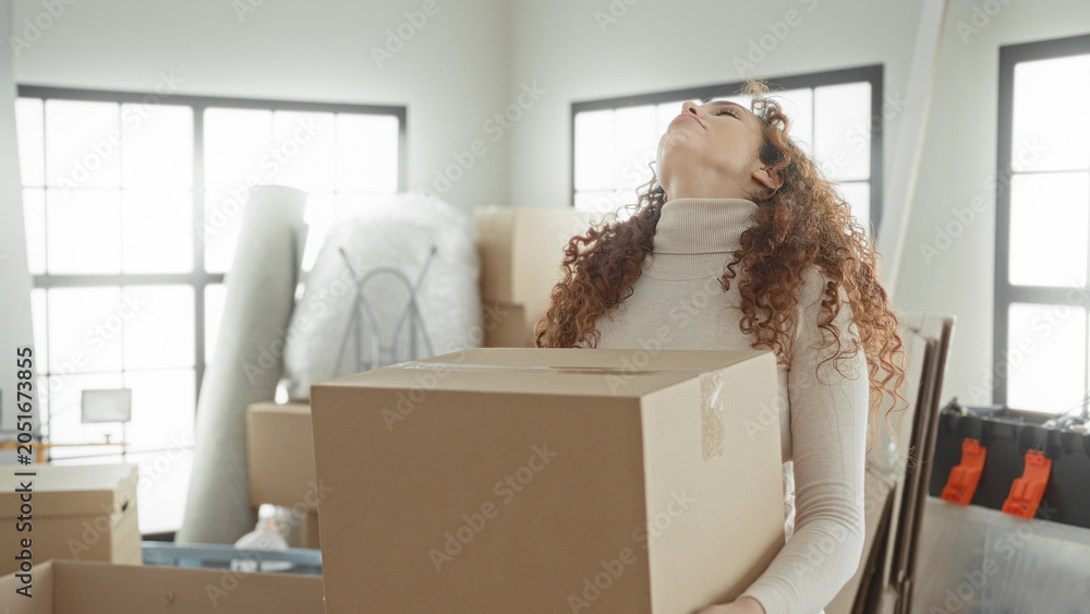 Fototapeta premium Woman carrying cardboard box with hands visible and grimacing, packing supplies stacked in apartment; moving stress.