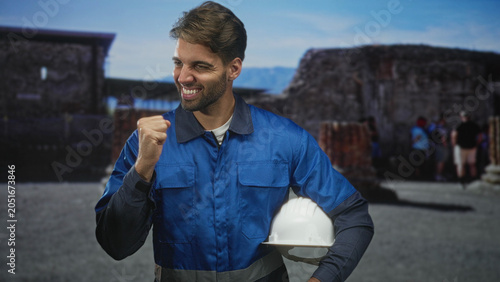 Quadro su tela Man construction worker wearing blue overalls, holding white hardhat with beard visible, smiling and fist pump in front of stone ruins building; pride duty