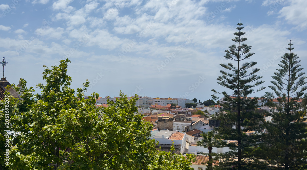 Fototapeta premium Tavira shows white houses and narrow streets from above, highlighting the tow's layout, greenery, and surrounding hills on a clear day in Algarve, Portugal