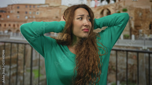 Foto Woman in green sweater covering ears with hands in building by roman ruins, grimacing against loud noise; annoyance noise stress