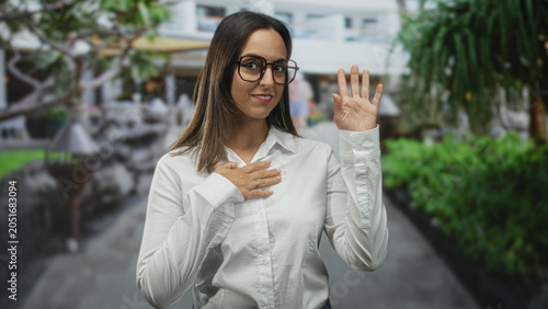 фотографія Woman wearing glasses and a white shirt places left hand on chest while raising her right hand in a wave on a street with outdoor seating and greenery; sincerity welcome