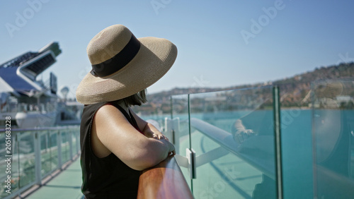 Fotografija Woman leaning on ship rail with bare shoulder visible, smiling while wearing sunhat and sunglasses on building deck walkway under clear blue sky; serenity vacation leisure