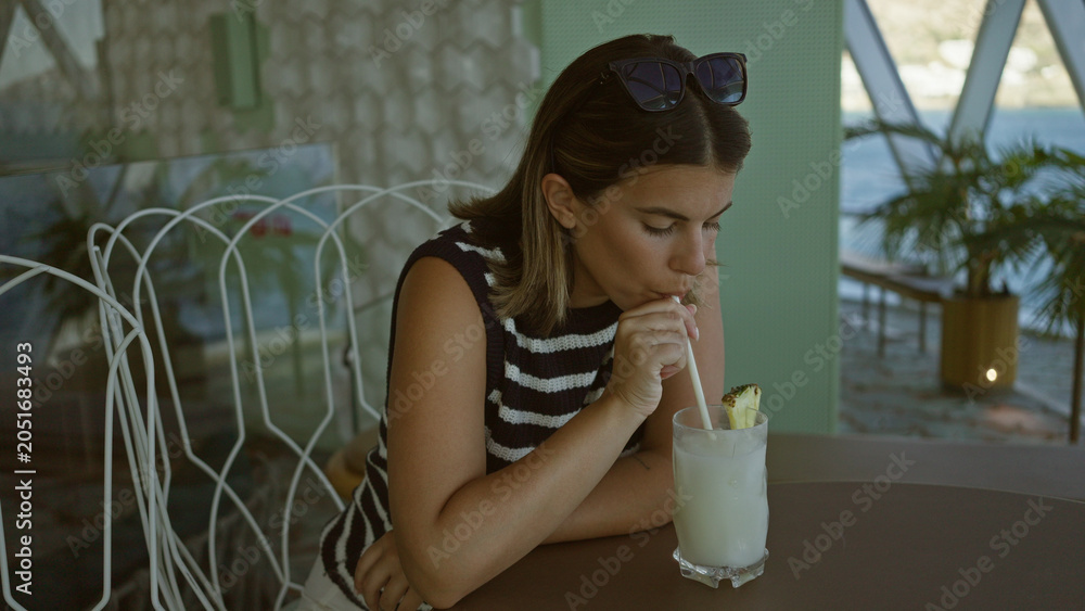 Obraz premium Woman stirs drink with straw at a table in a building by the water, sunglasses on head; contemplation moment.