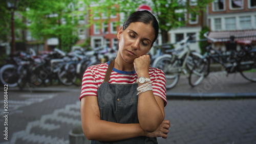 Obraz na plátne Woman with hand on cheek and crossed arms wearing apron standing on street beside parked bicycles; pensive reflection