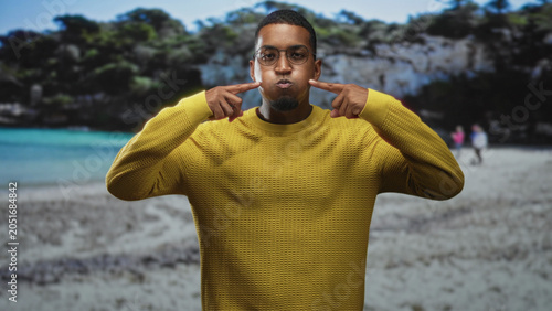 Slika na platnu Young black man with glasses puffs cheeks and points to face on a sandy beach in a yellow sweater; playful expression