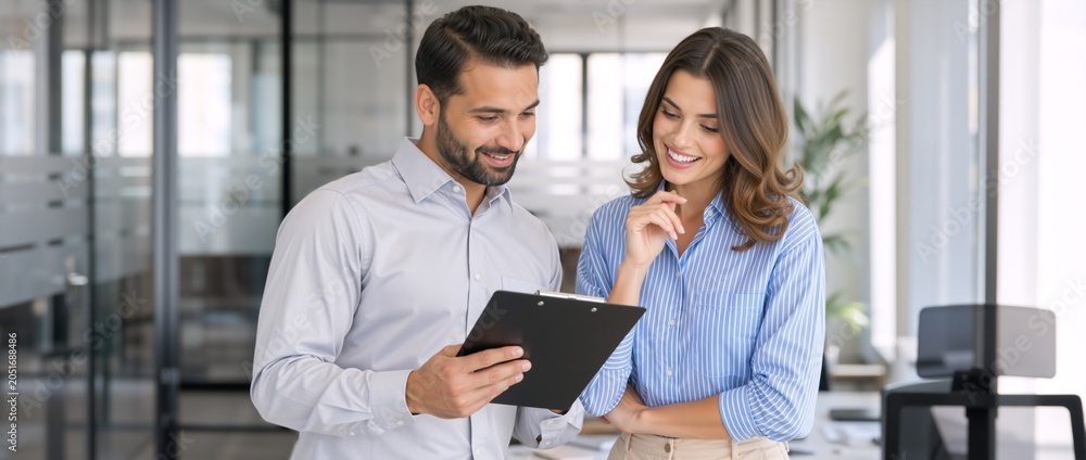 Fototapeta premium Happy business colleagues reviewing a document on a clipboard in a modern office. Professional man and woman discussing a project. Positive teamwork