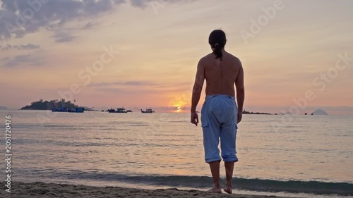 Rear view of a shirtless man standing on a sandy beach at sunset, facing calm water with distant boats, soft waves, and a pastel sky