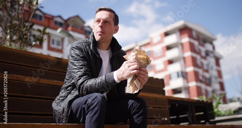 A man relaxes on wooden steps, savoring his lunch while enjoying bright sunshine and the lively atmosphere of the city. Surroundings feature modern buildings and clear skies