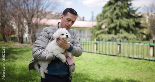 In a peaceful park filled with greenery, a man cradles a fluffy white dog in his arms, surrounded by trees and a clear blue sky, enjoying a moment of companionship and tranquility