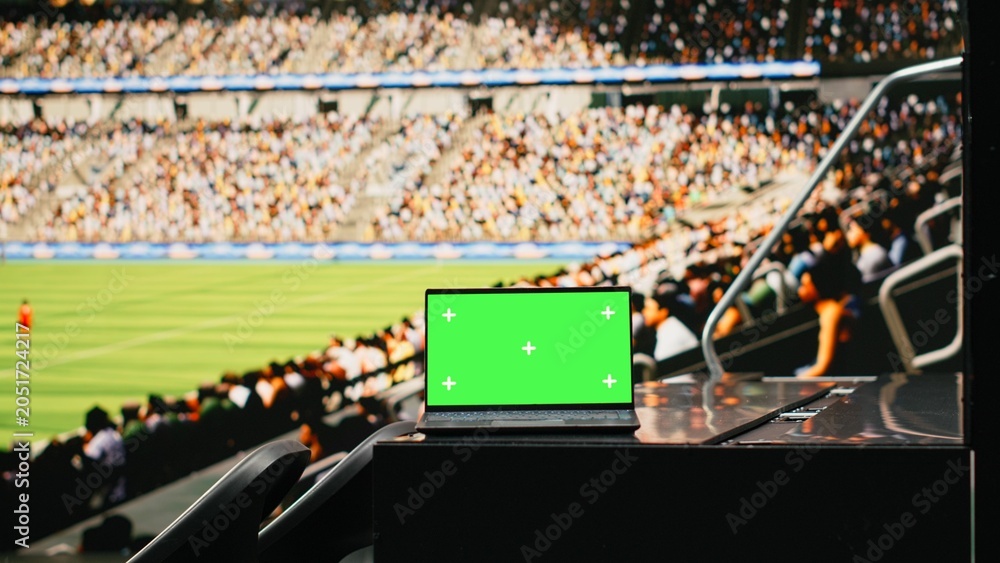 Naklejka premium Mockup green screen laptop in a crowded stadium during football game, fans cheering in tribunes under dramatic stadium lights with football pitch and packed audience visible in background.